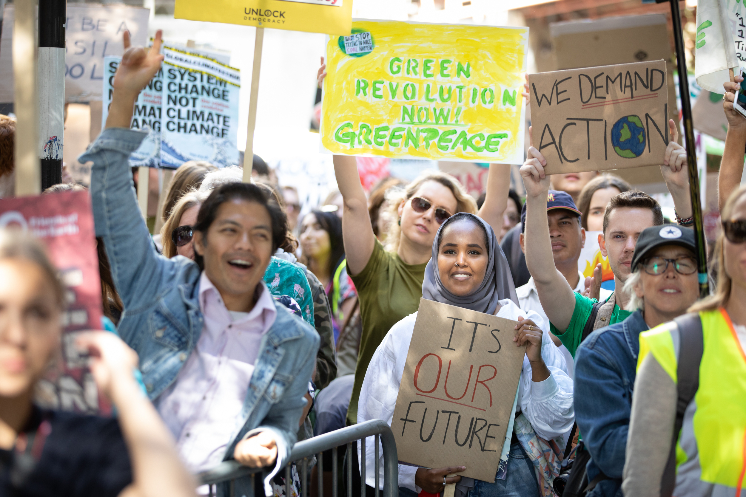In bright sunshine, a smiling crowd of people hold up signs calling for a greener world. One protestor in the front row raises his fist in the air triumphantly.