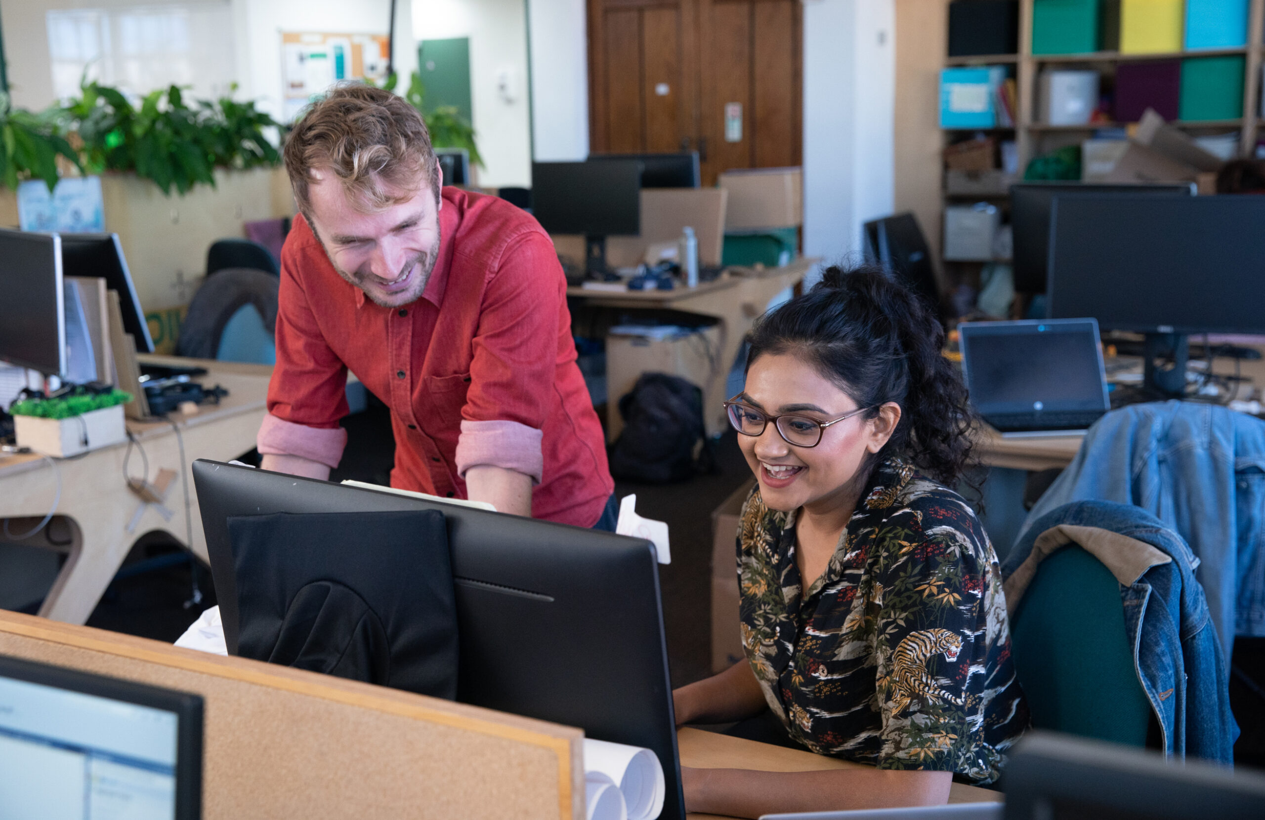 Two people of different genders and races smiling together looking at a computer screen in an office full of desks and plants