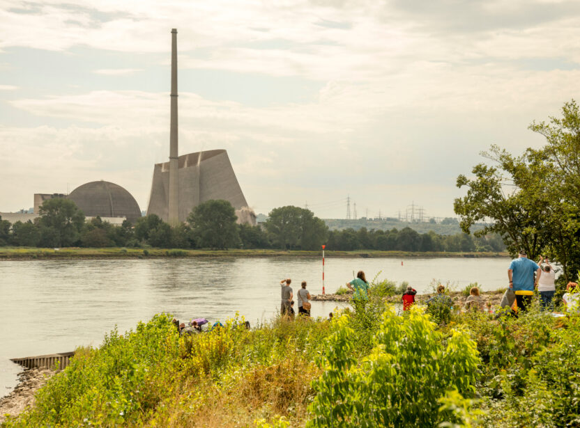 People play in lush vegetation by a river. On the opposite bank sits a large nuclear power station in the process of being demolished.