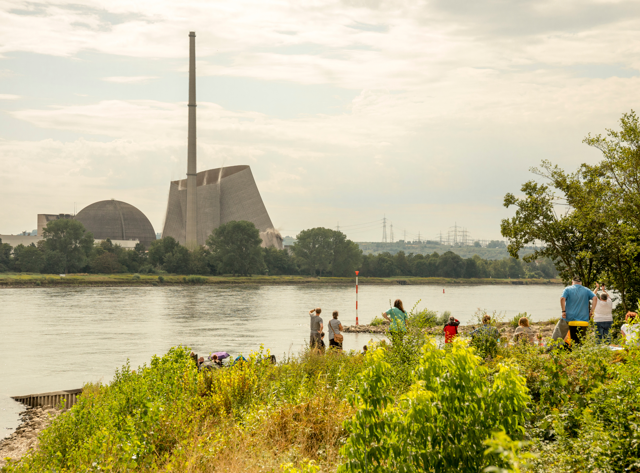 People play in lush vegetation by a river. On the opposite bank sits a large nuclear power station in the process of being demolished.