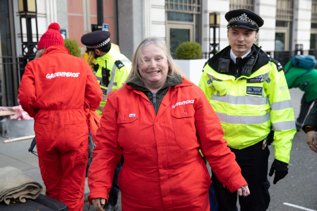 Activist in a boiler suit smiles into the camera as she's led away by a police officer
