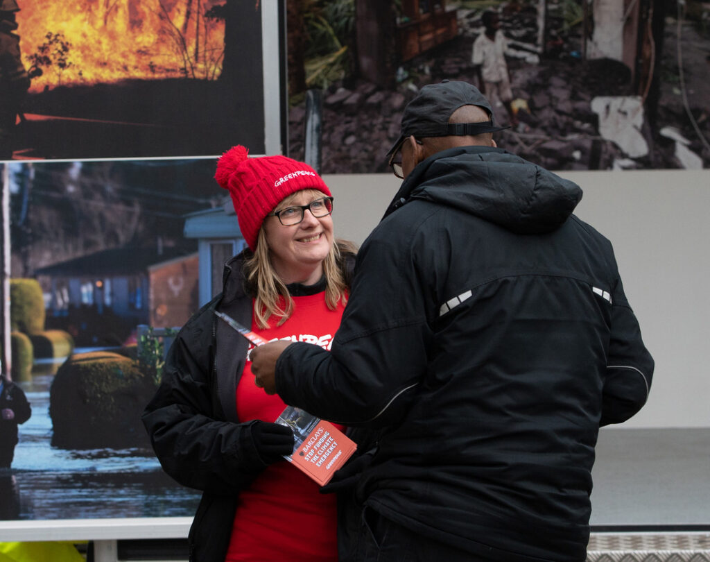 An activist wearing a Greenpeace branded bobble hat smiles at a member of the public as they have a conversation during an action.