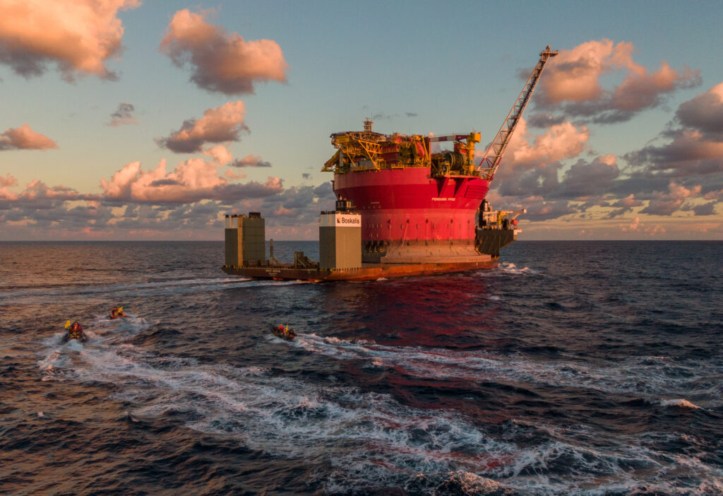 Giant red cylindrical oil platform travels on a ship along the ocean. Activist boats zoom towards it. It looks like David approaching Goliath because they're so small in comparison.