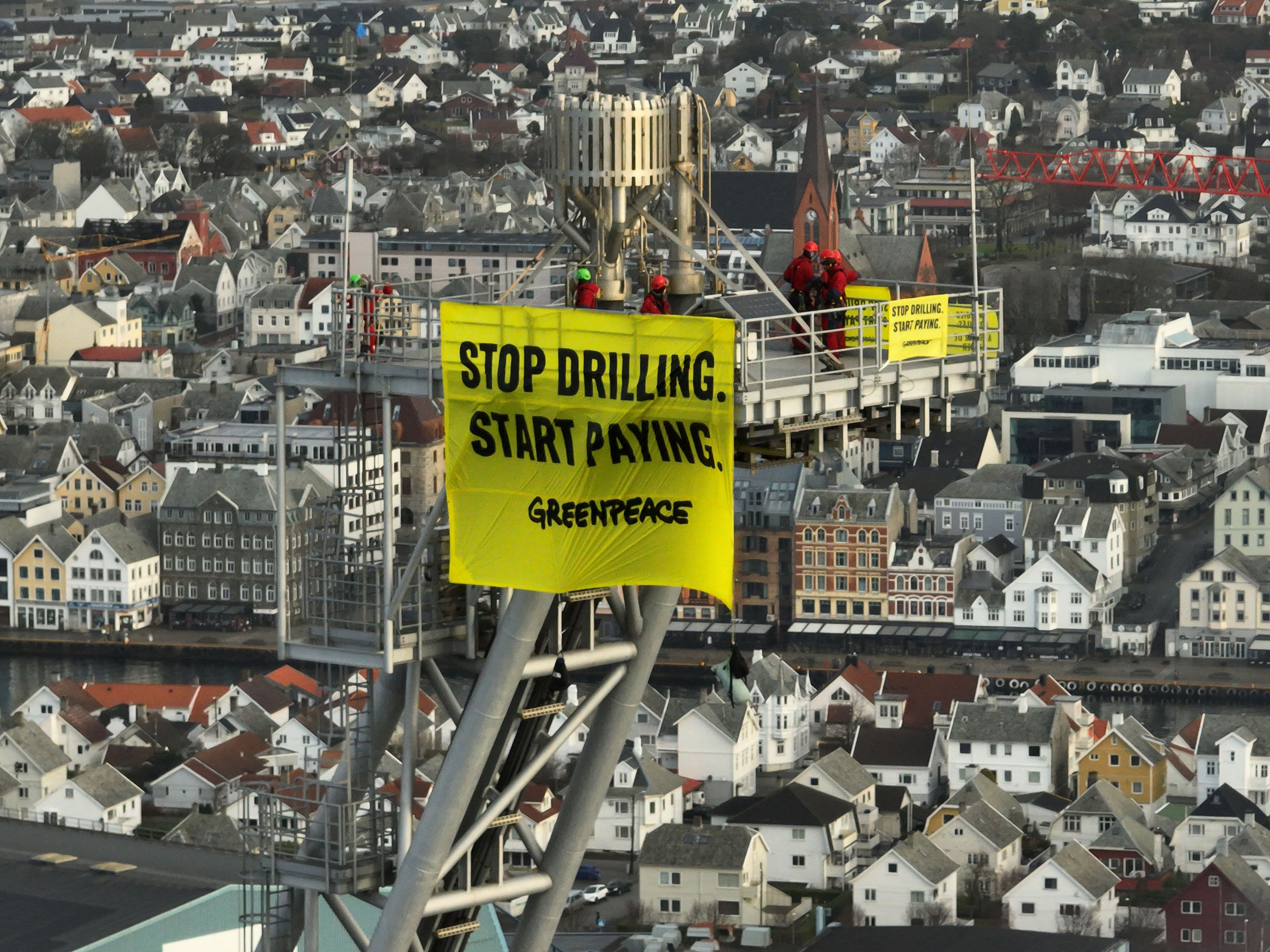On a background of dense greyscale cityscape filling the entire frame, protestors are holding a yellow banner reading ‘Stop drilling. Start Paying. Greenpeace' high above on a metal frame