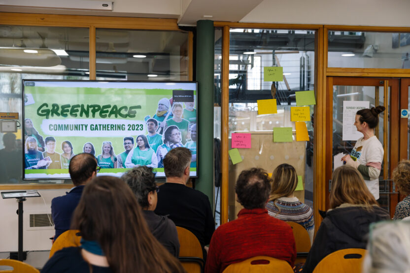 A crowd of people from beind, they are sat looking at a speaker in front of a galls wall with post-its and a screen reading 