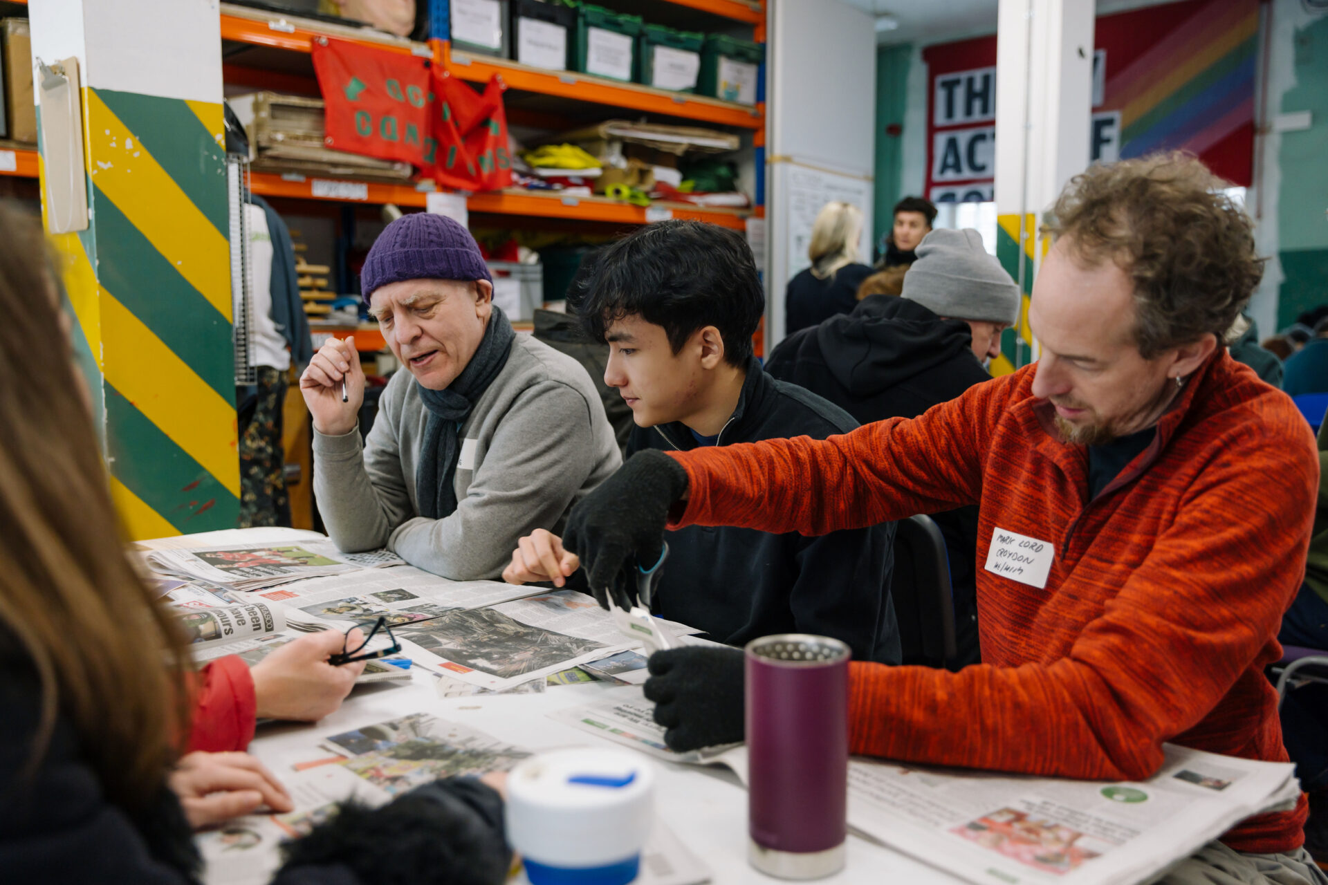 Three men of different ages sit at a table, looking at a spread of newspapers, in a colourful warehouse with shelves and banners visible in the backgrounud