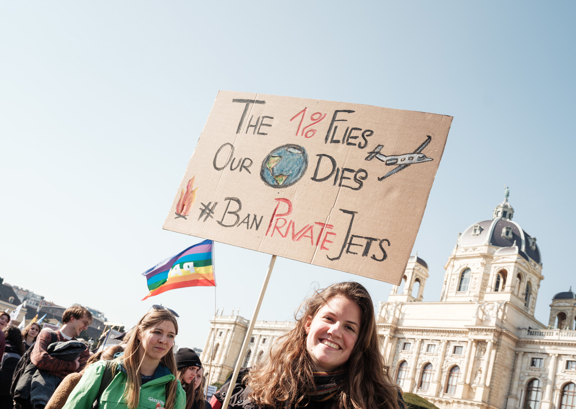 Smiling protestor holds a handwritten sign saying 'The 1% flies, our planet dies. Ban private jets.'