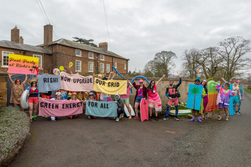 A colourful parade of people wearing shirts, swimming caps, bikinis, wetsuits and swimming gear like snorkels, caps, goggles and tropical accessories like straw hats. They are displaying a boards with a plam tree painted on it, one with a sunset colours background and palm tree icons, with handpainted text reading 