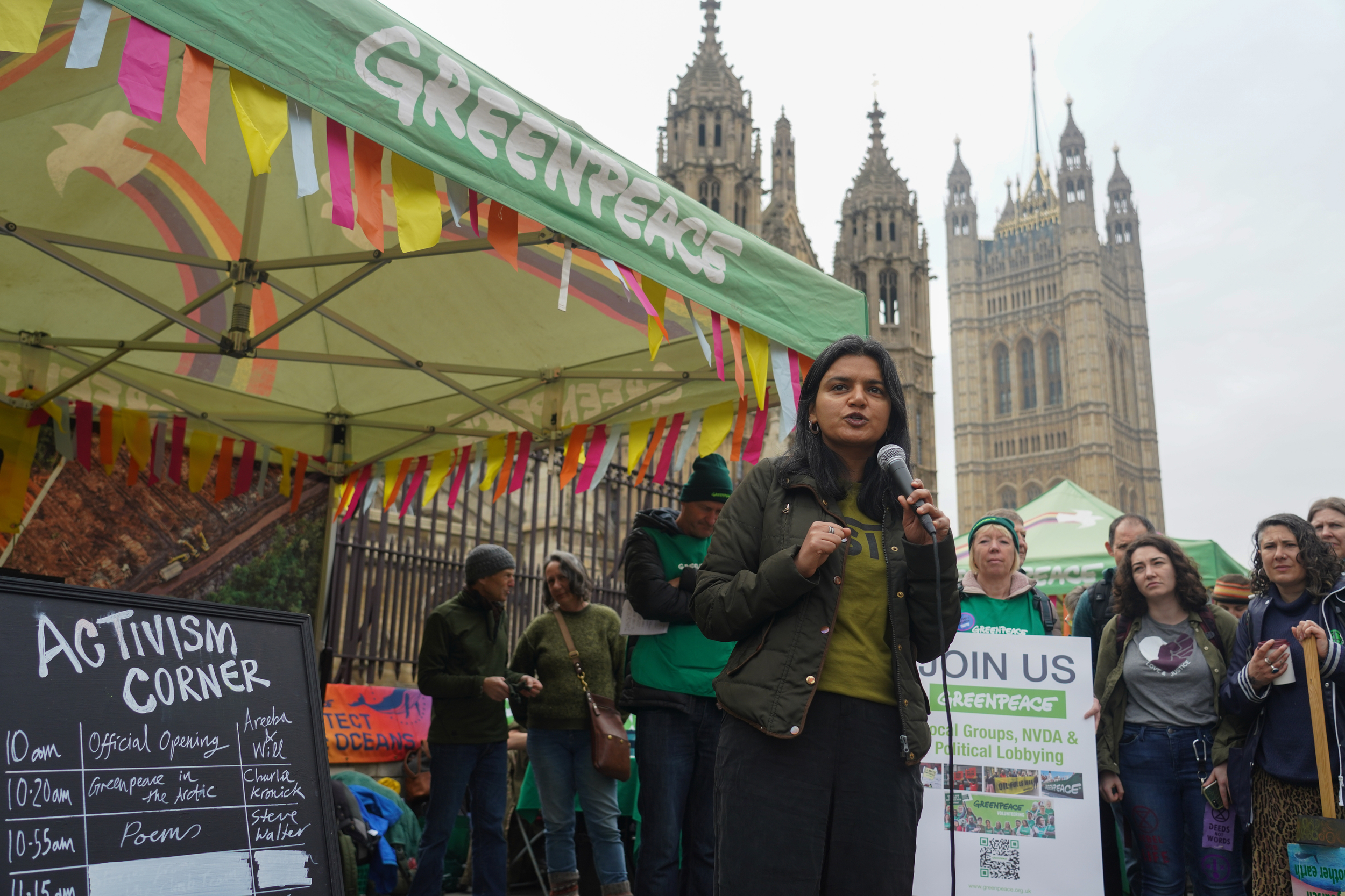 Areeba Hamid speaks into a microphone outside a Greenpeace branded gazebo, with UK Parliament in the background.