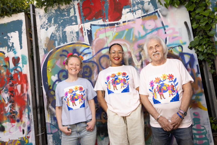 Three people wearing tshirts with the same colourful design stand in front of a wall covered in graffiti. They're posing informally and smiling into the camera.