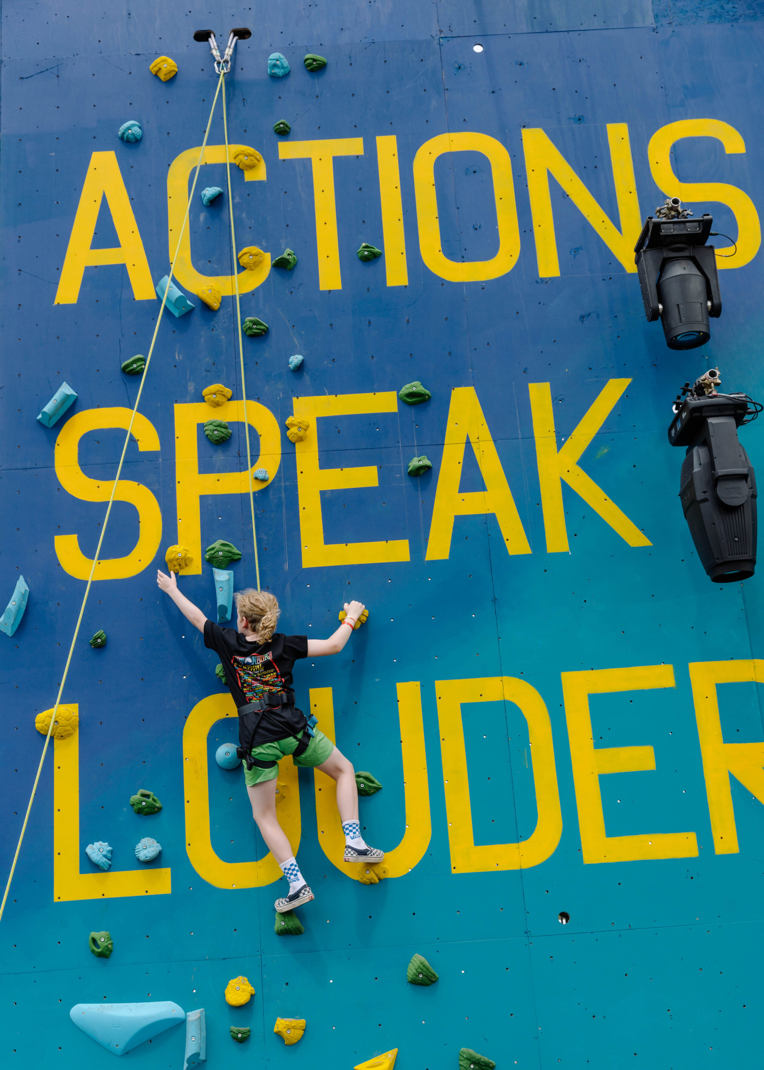 Climber climbs up a climbing wall painted with a giant 'actions speak louder' slogan in the Greenpeace field at Glastonbury.
