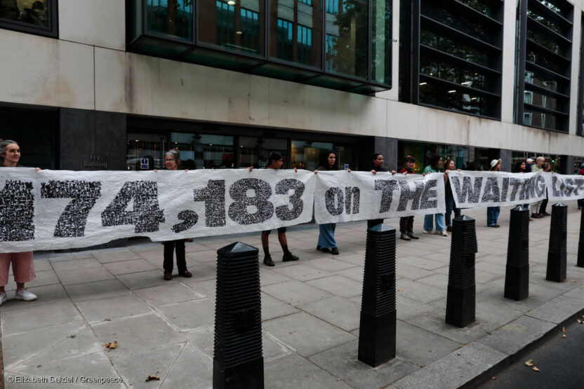 A 30 metre banner is held open by volunteers on a pavement outside an office. The banner reads 