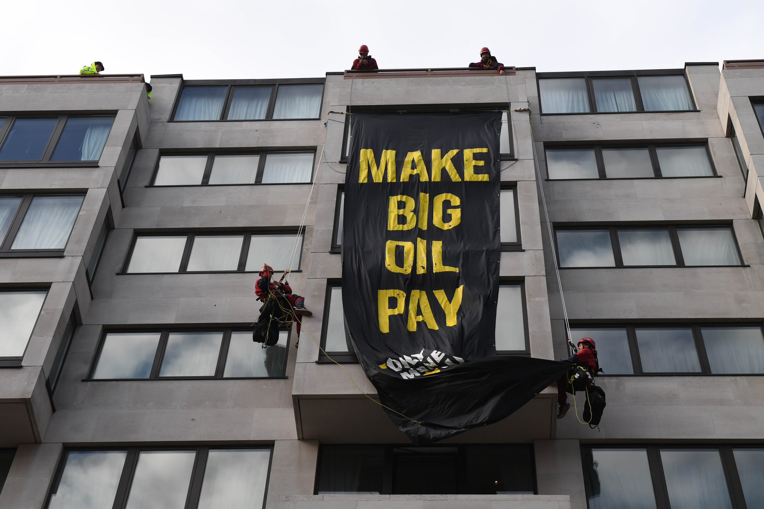 Climbers on a building unveil a big black banner with yellow text. The banner reads: Make big oil pay