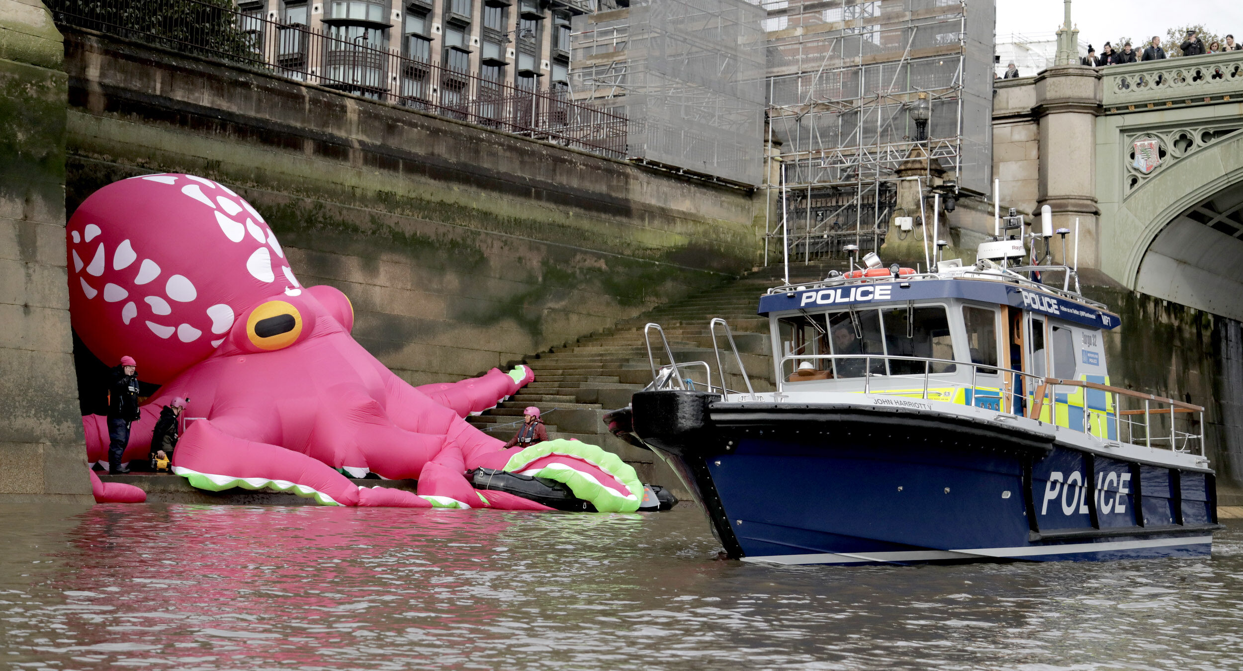 A giant pink inflatable octopus sits on the bank of the Thames directly below the Houses of Parliament, with a police boat alongside