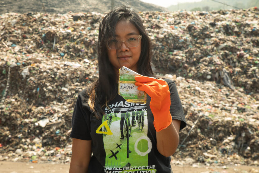 An activist in a Greenpeace t-shirt holds up a piece of plastic litter with Dove branding. There's a huge pile of plastic waste in the background.