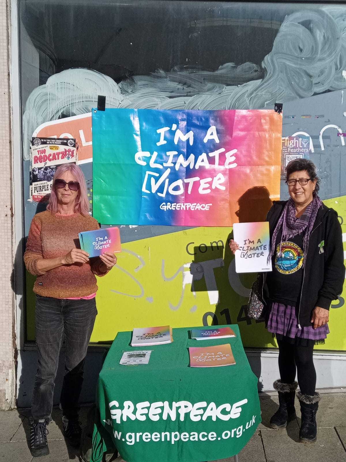 Two women of different ethnicities stand in front of a disused shop holding flyer and posters standing in front of a large colourful "I'm a climate voter" banner, and a table with a green Greenpeace tablecloth