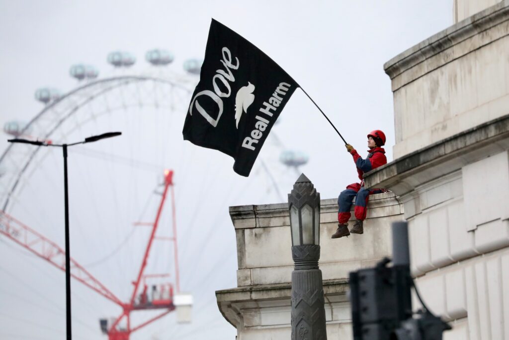 An activist in a helmet and boiler suit sits on a ledge of an ornate stone building, waving a black flag saying 'Dove - Real Harm'. The London Eye is visible in the background.