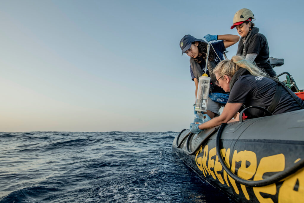 Crew members lean over the side of a Greenpeace-branded inflatable boat to retrieve sampling equipment from the water.