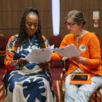 Two women of different ethnicities sit side by side comparing notes in a conference room. one has a "I'm a Climate Voter" sticker on her cardigan.
