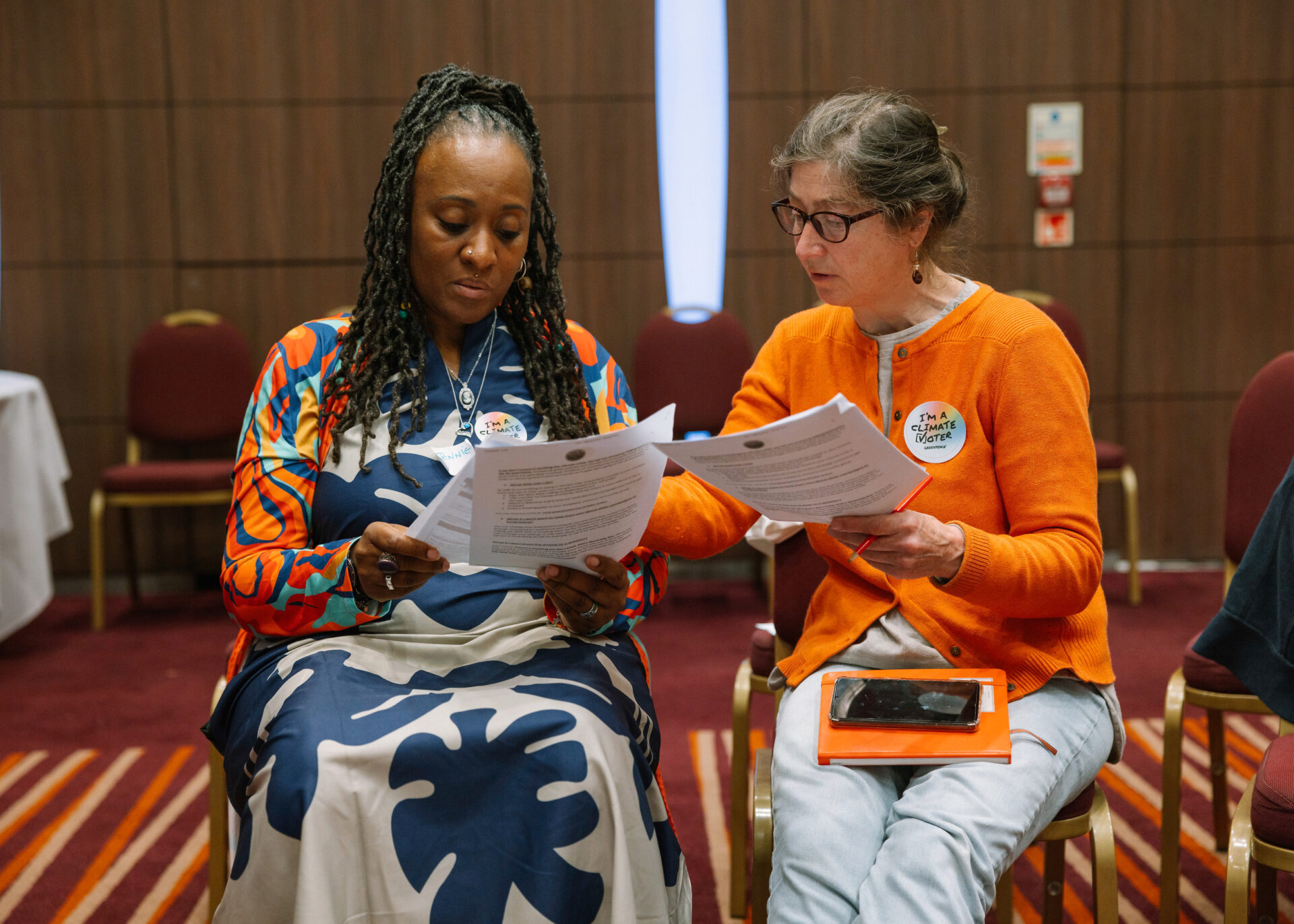 Two women of different ethnicities sit side by side comparing notes in a conference room. one has a "I'm a Climate Voter" sticker on her cardigan.
