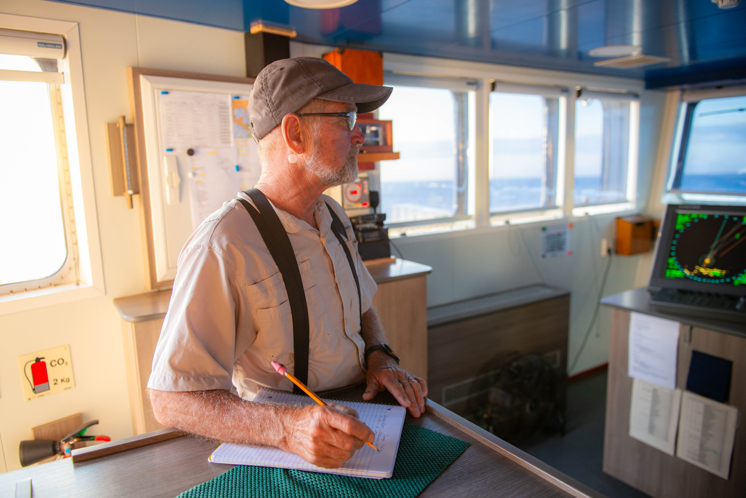 A man in a cap, glasses and suspenders stands in a ship's cabin writing something on a clipboard