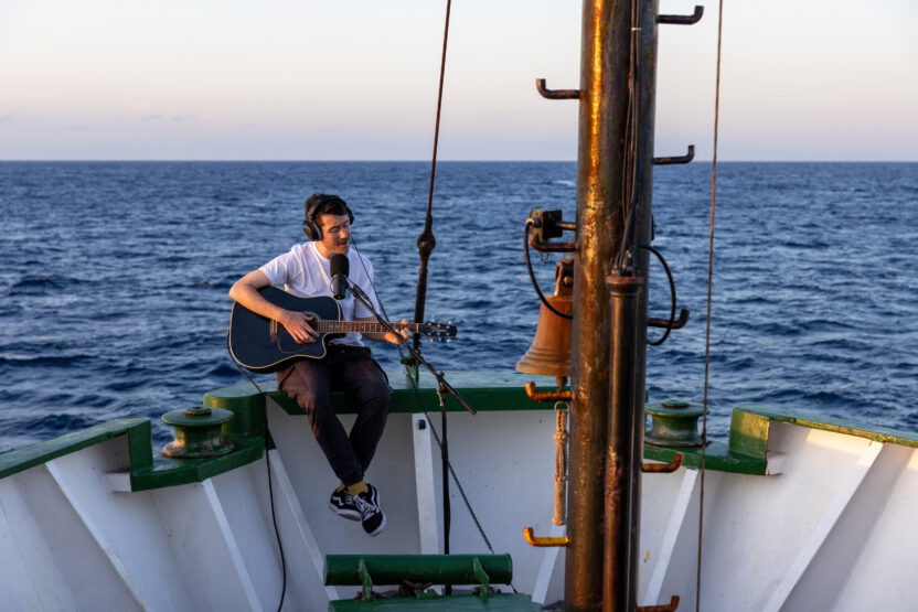 Dan Smith from Bastille sits on bow of a ship playing an electric guitar and singing into a microphone with open ocean behind him