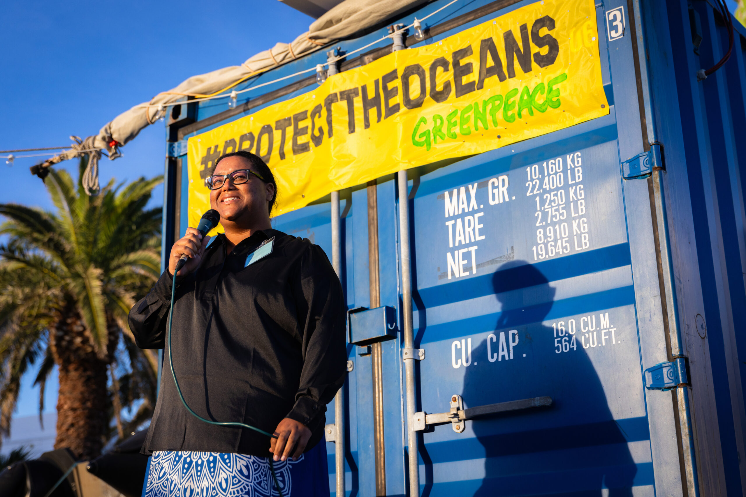A woman of colour stands smiling and holding a microphone in front of a blue metal container, with a yellow Greenpeace banner reading "Protect the Oceans" behind her
