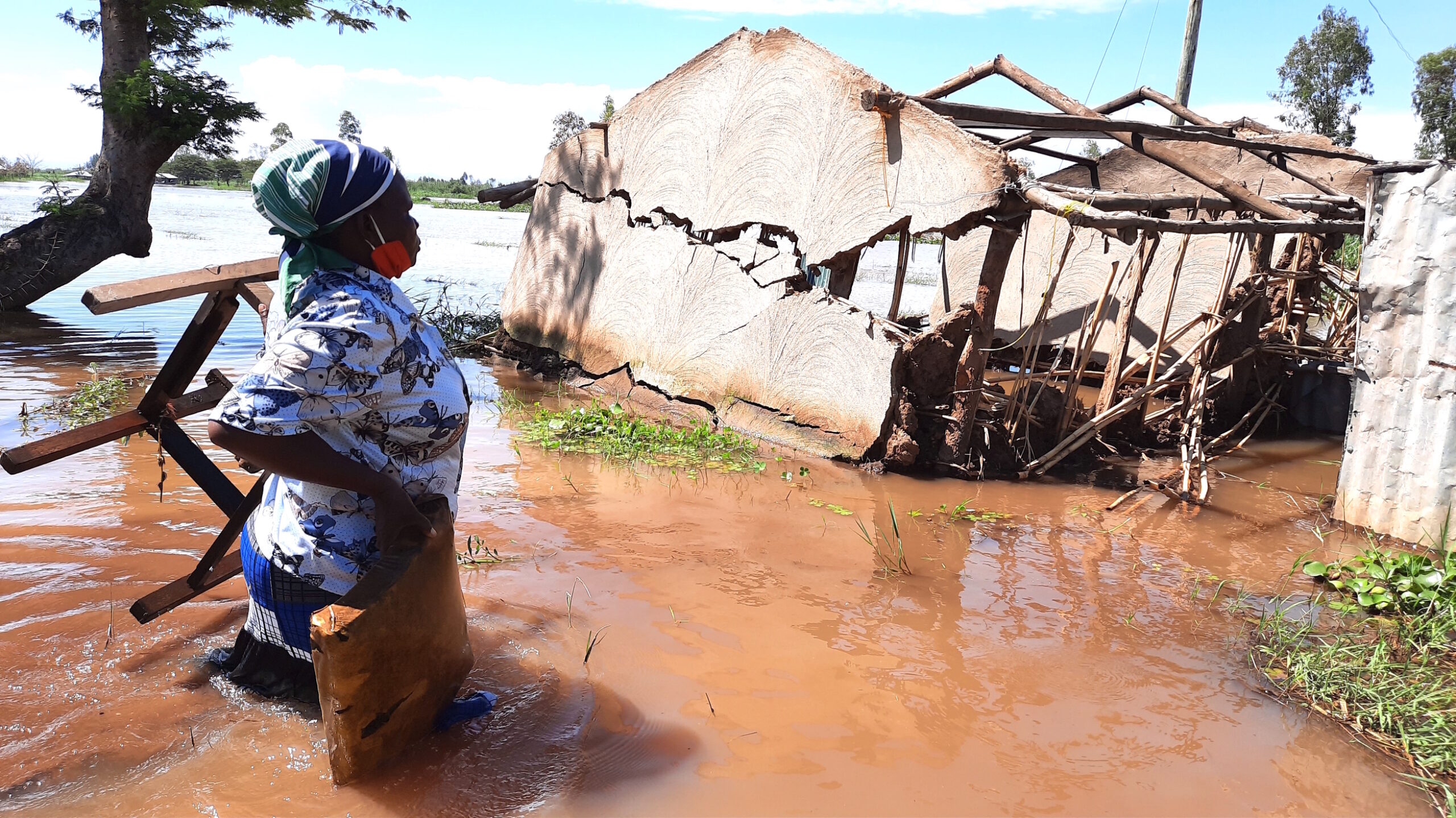 A Black person carries furniture through muddy, waist-deep flood water towards a wooden-framed building that's been severely damaged.