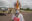 Person in a traditional Indigenous headdress and facemask stand in front of a light aeroplane on a runway