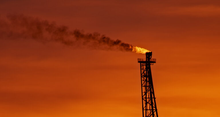 At the top of a metal gantry, a bright flame gives off black smoke, blowing horizontally across a deep orange sunset sky.