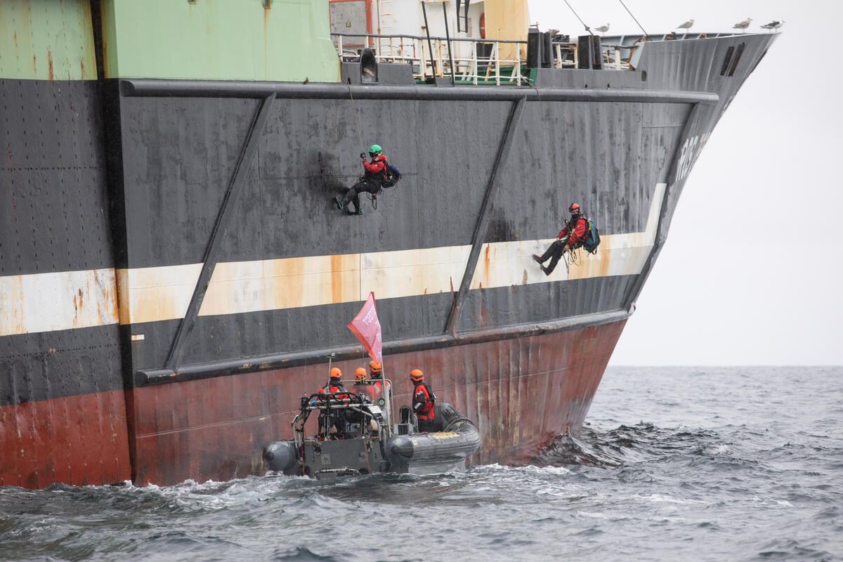 Two activists hang from the side of a large fishing ship. A small Greenpeace boat drives alongside.