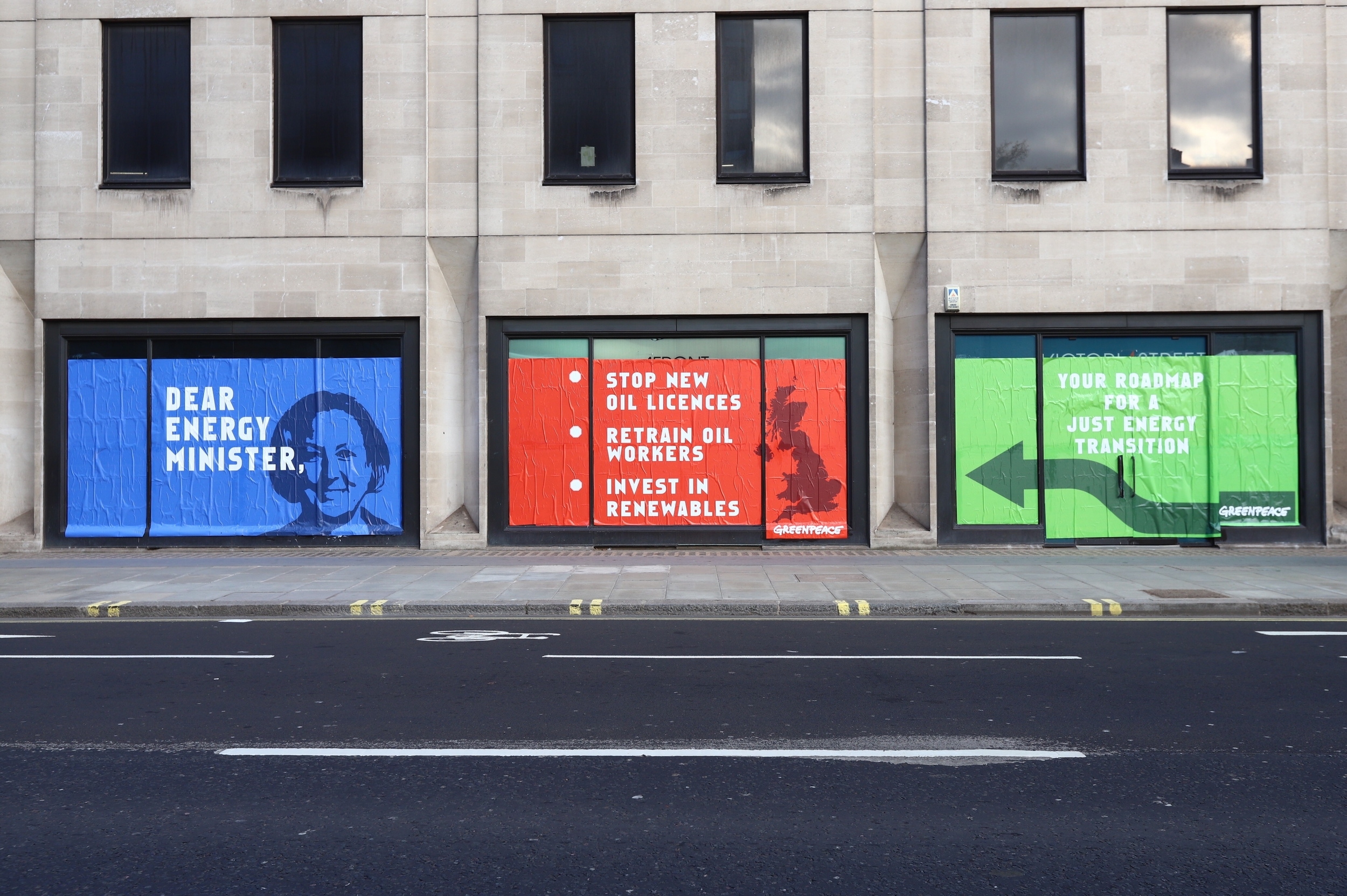 A building with three full-height, long windows plastered with a message to the energy minister. The blue, red and green posters have the energy minister's face, a map of the UK and an arrow on them, and read 'Dear energy minister. Stop new oil licenses. Retrain oil workers. Invest in renewables and the final green poster with the arrow points to the previous list, reading 'Your Roadmap for a Just Energy Transition'