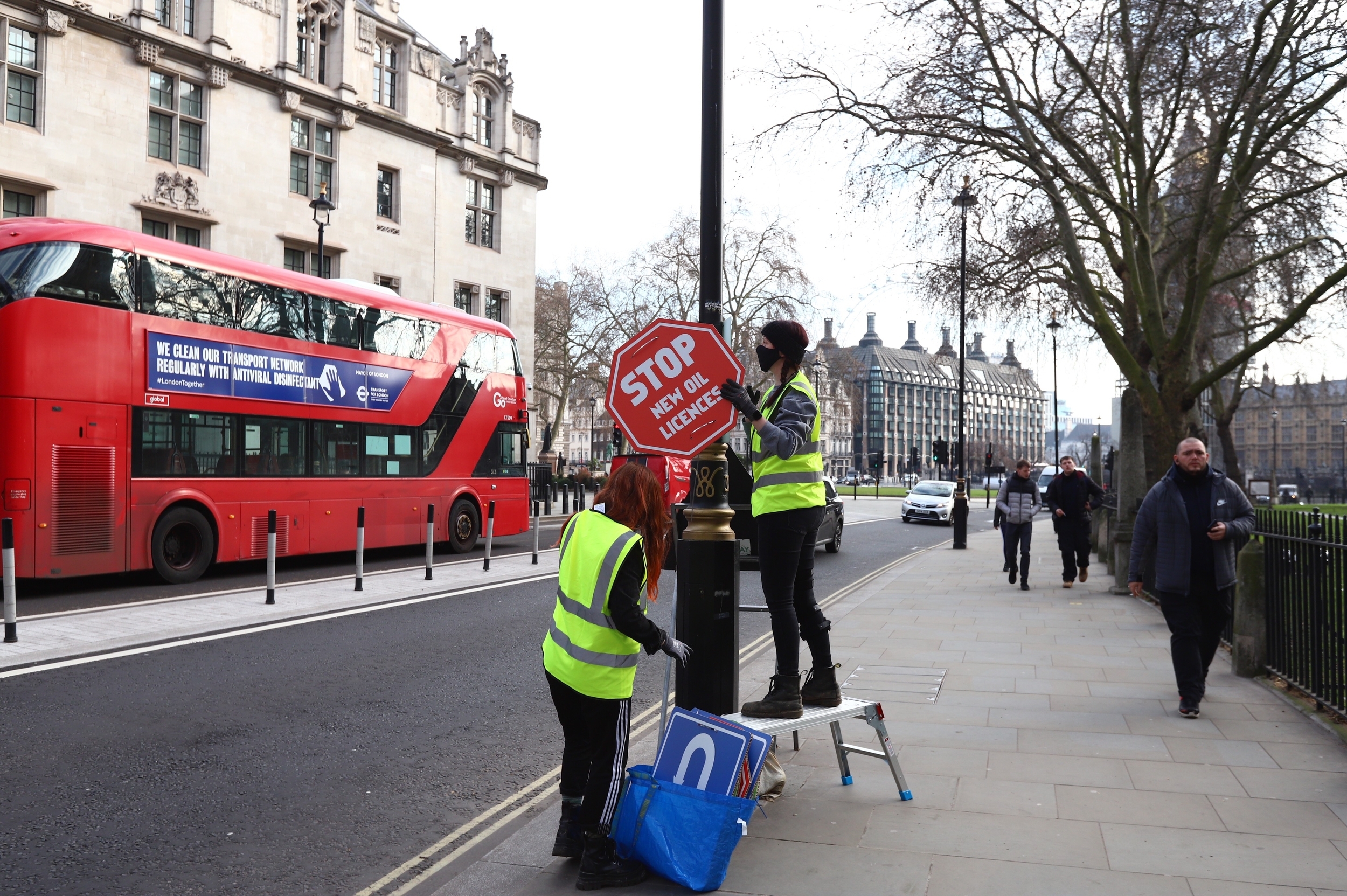 Two women in hi-vis mounting signage on a lamp post. The sign is red and shaped like a stop sign, reading Stop new oil licenses. A red London bus and Parliament Square are in the background.