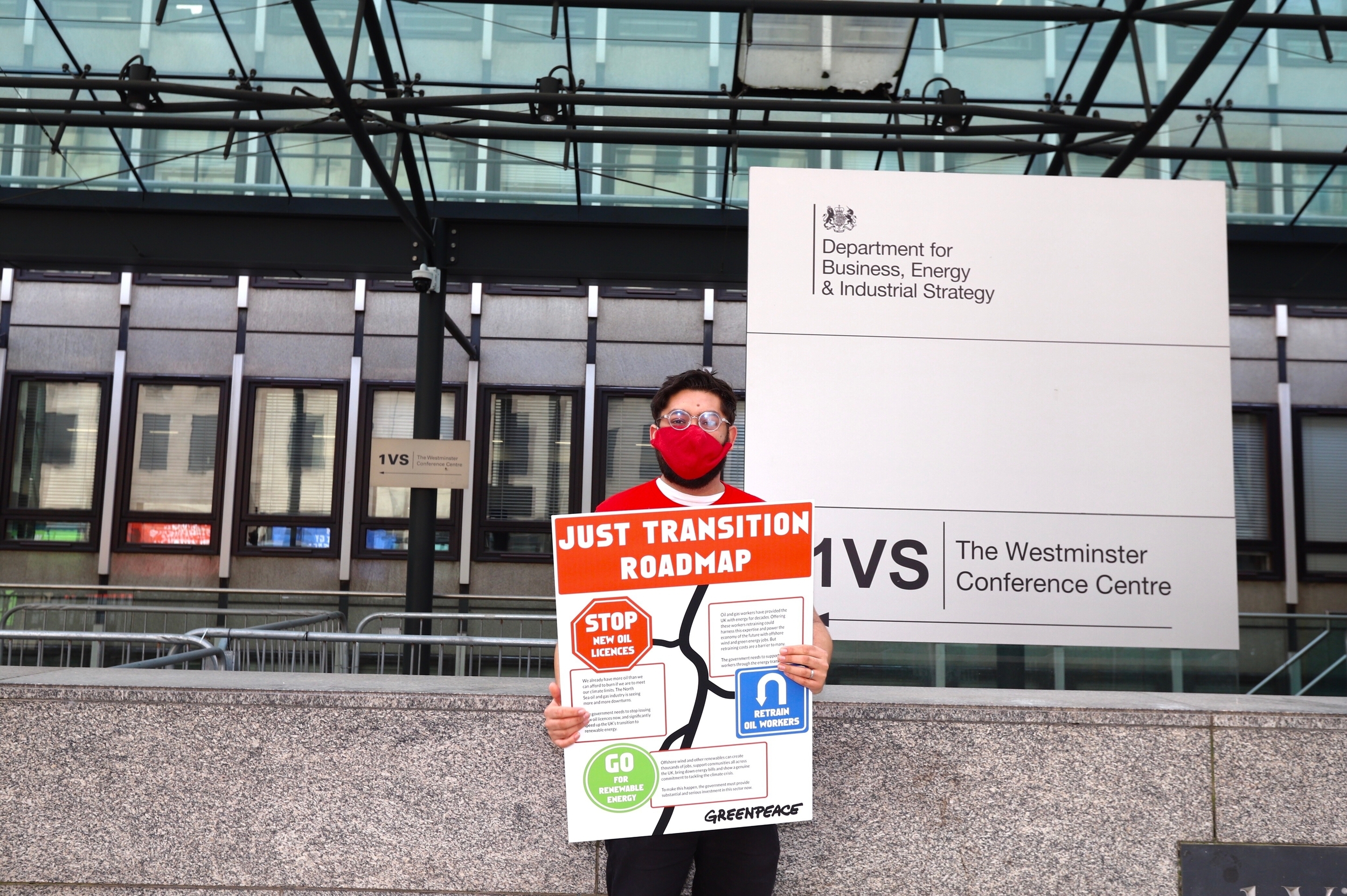 A man stands outside the BEIS department, holding a poster reading 'Just transition roadmap' with 'Stop new oil licenses' and 'Invest in renewable energy' on signs that mimic road signs, with arrows in between.'
