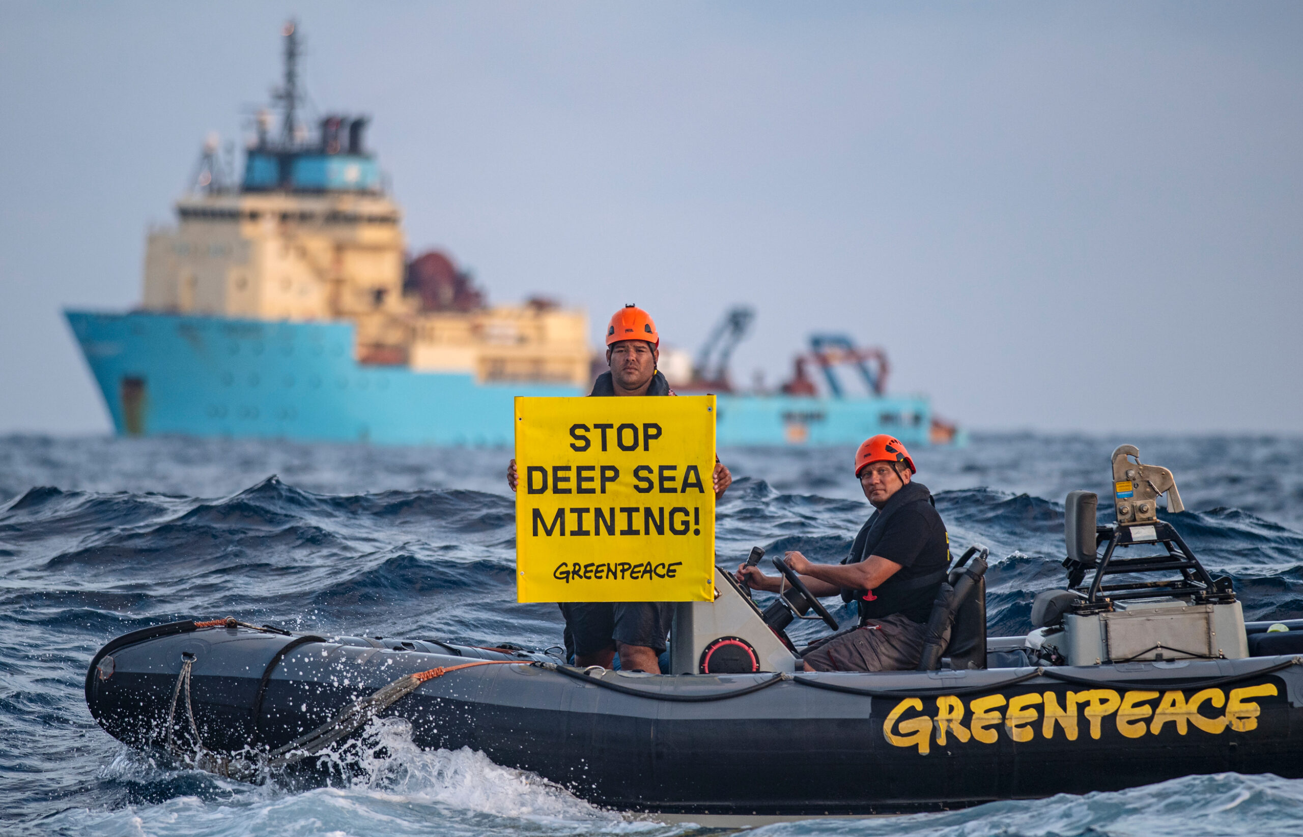 Two Greenpeace activists in an inflatable boat on the ocean, with a big ship in the background. One stands holding a a banner that says "Stop deep sea mining", while the other drives the boat.