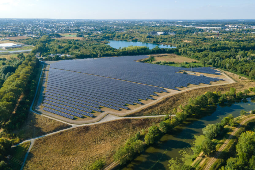 An aerial shot of a large field of solar panels, surrounded by trees and a small lake, with some houses and other neighbourhood buildings dotted across the horizon in the distance