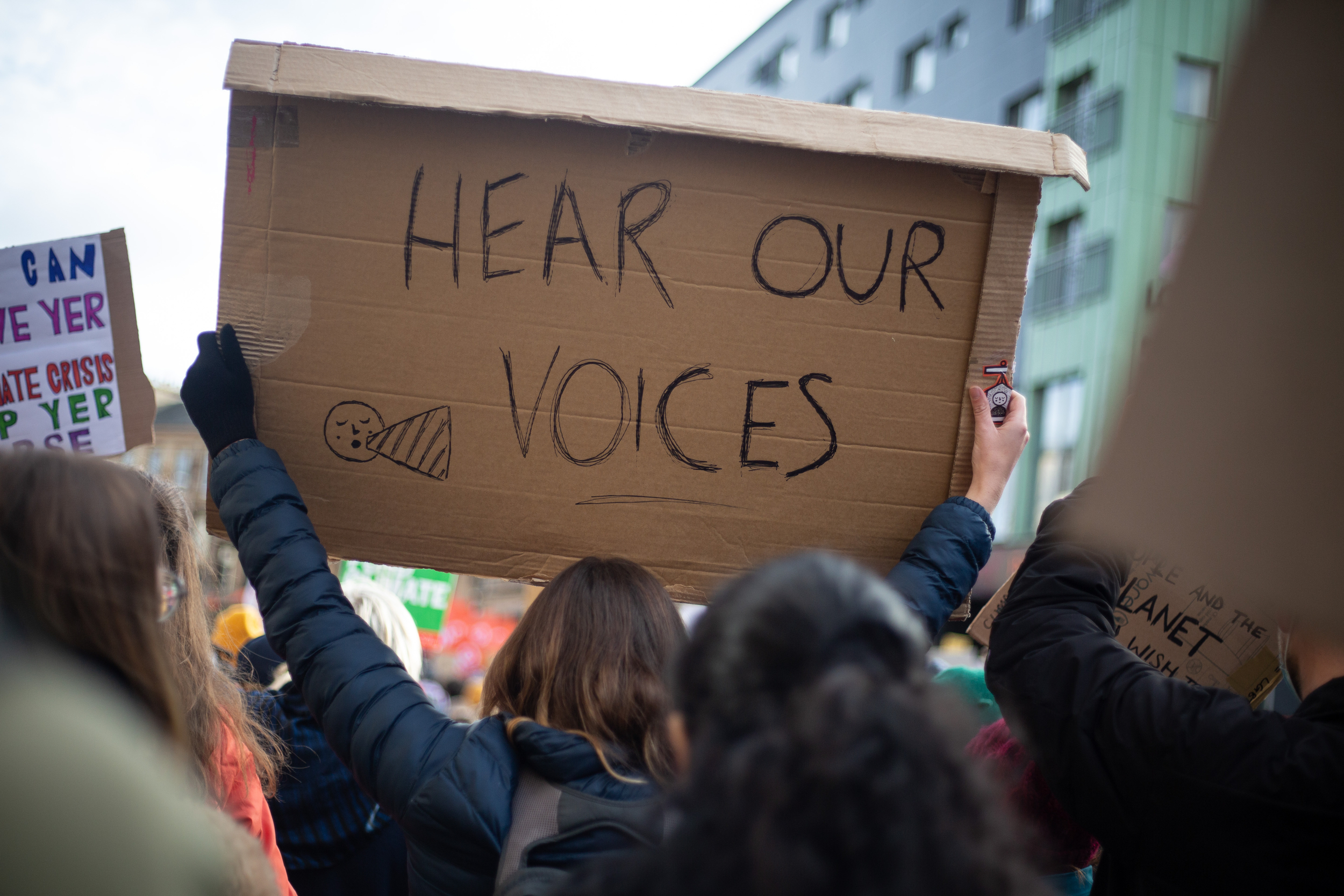 Protester viewed from behind, holding up a handwritten cardboard placard that says 'hear our voices'.