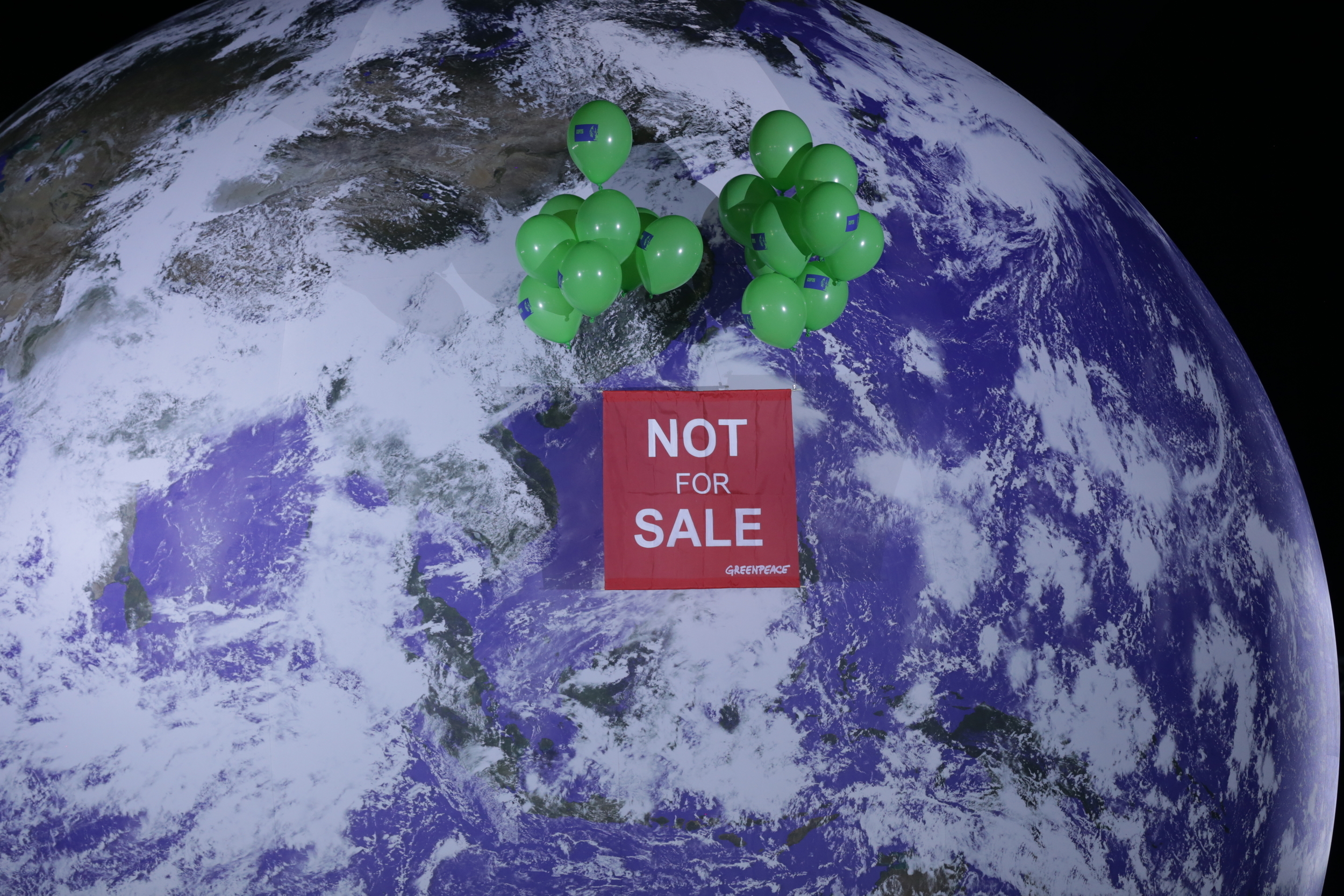 Lifted by green balloons, a red banner floats in front of a giant realistic model globe reading 'Not for sale'.