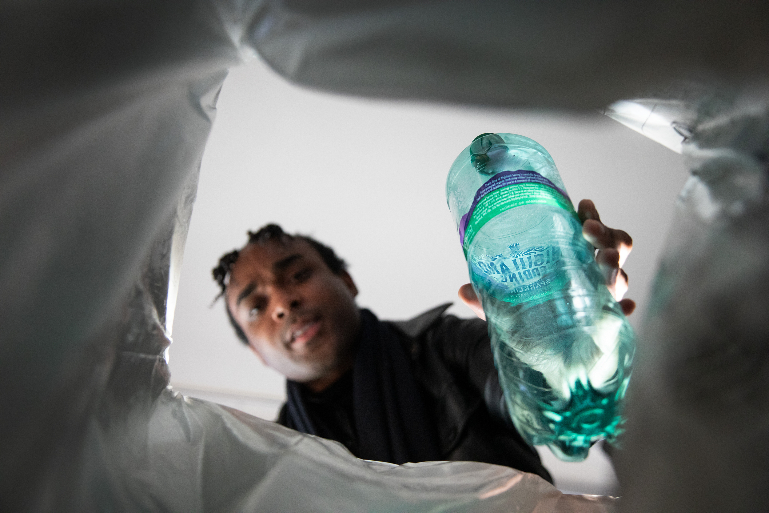 View from inside a bin, a person putting a green plastic bottle into it, framed by the plastic bag lining the bin.