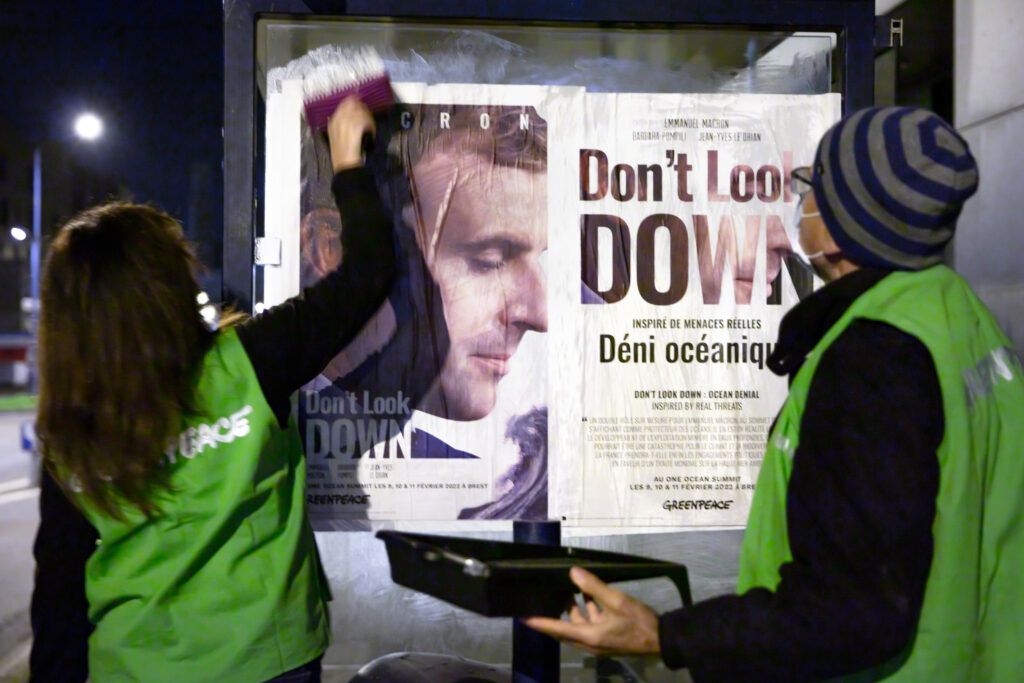Two Greenpeace activists (one woman with long hair and one with a warm stripey hat), both in bright green tabards mount a backlit poster in an advertising frame. It is a picture of Macron with newspaper style page reading 'Don't Look Down'