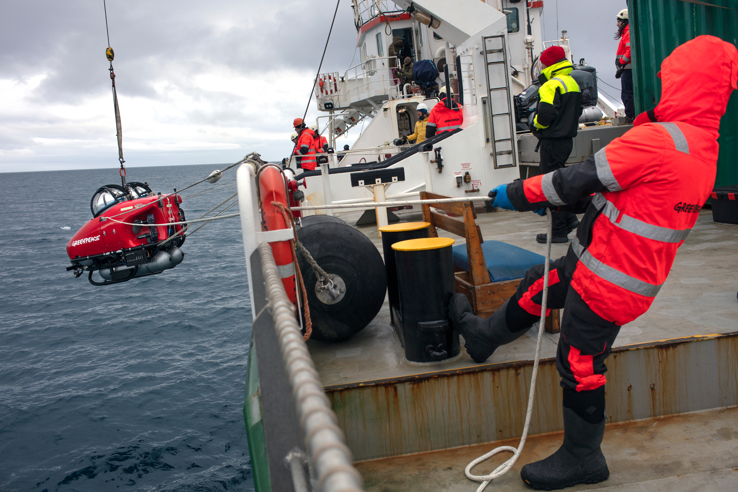 Ship crew hold ropes stabilising a small red submarine as it's lowered into the water from the ship's deck