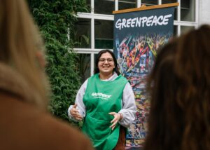 A speaker in a green tabard gesticulates as she delivers a talk in front of a Greenpeace banner.