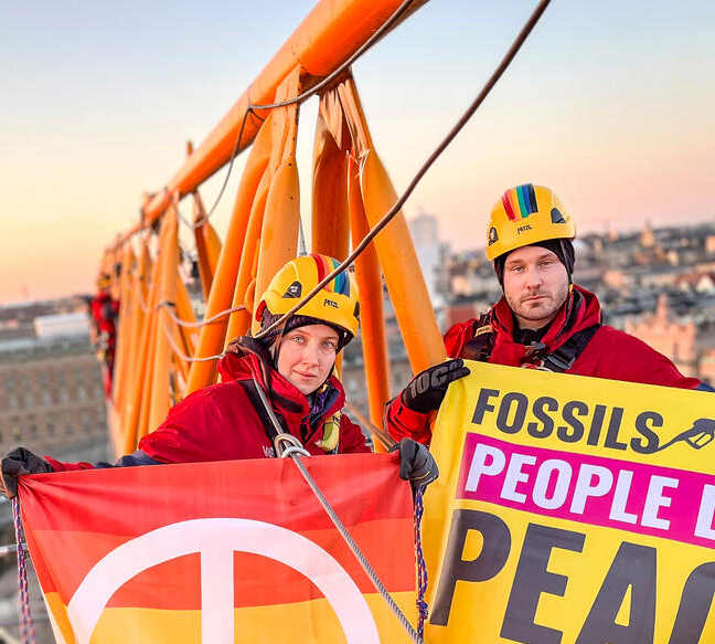 Two climbers wearing helmets and red overalls hold campaign banners on the arm of a crane, high above a beautiful urban sunrise.