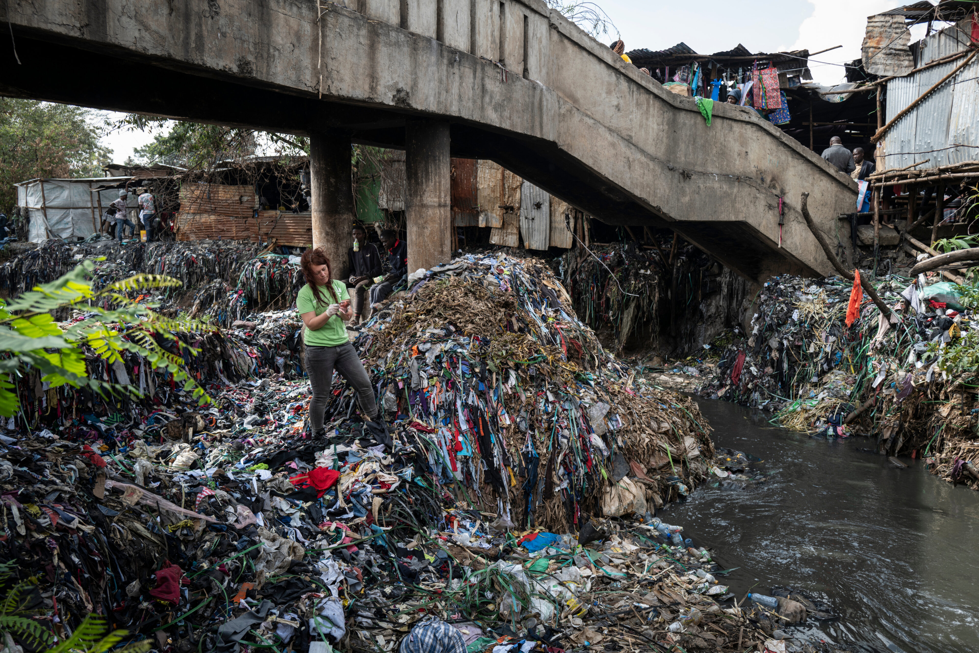 A woman in a green t-shirt and rubber gloves stands in a narrow waterway that's choked with plastic pollution piled almost head high in the water