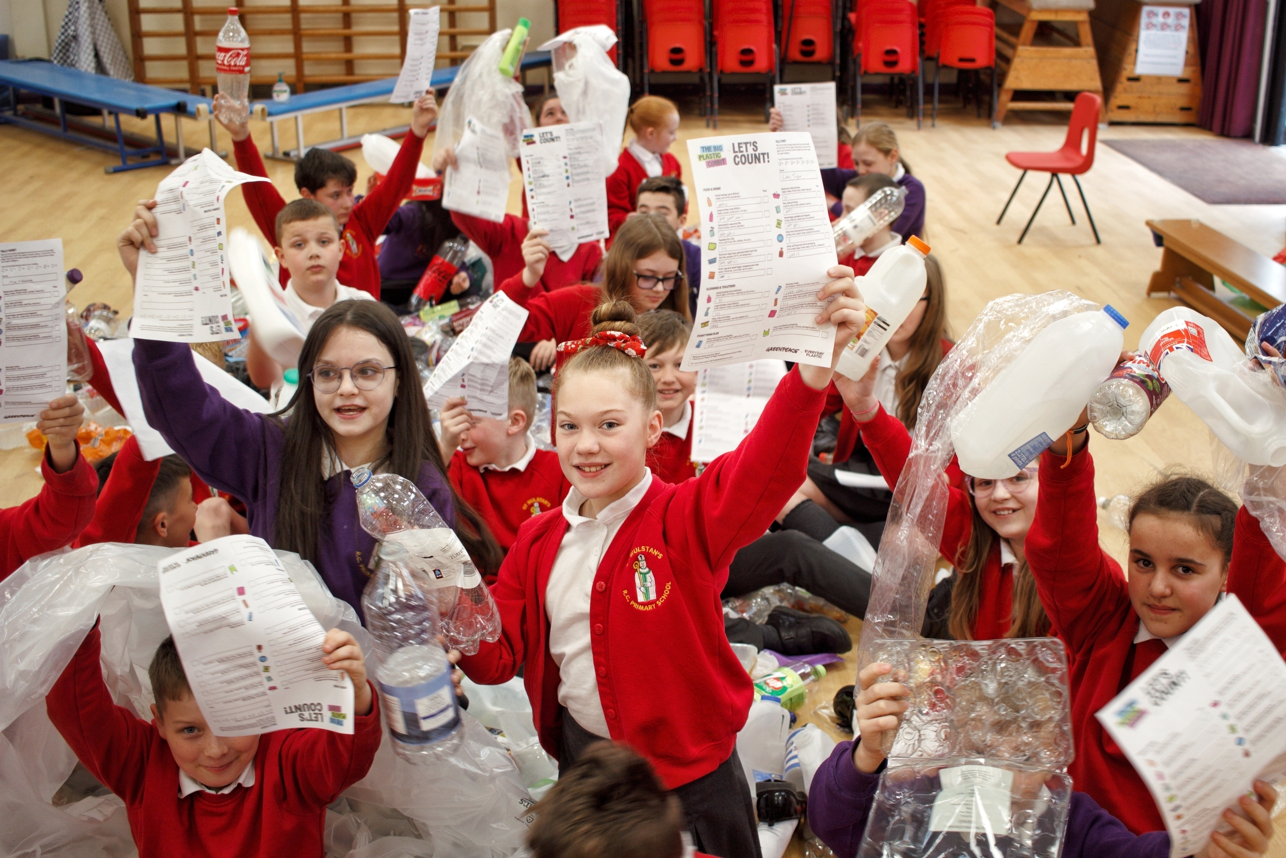 Primary school children sit amongst empty plastic containers holding up tally sheets for counting the different types of plastic.