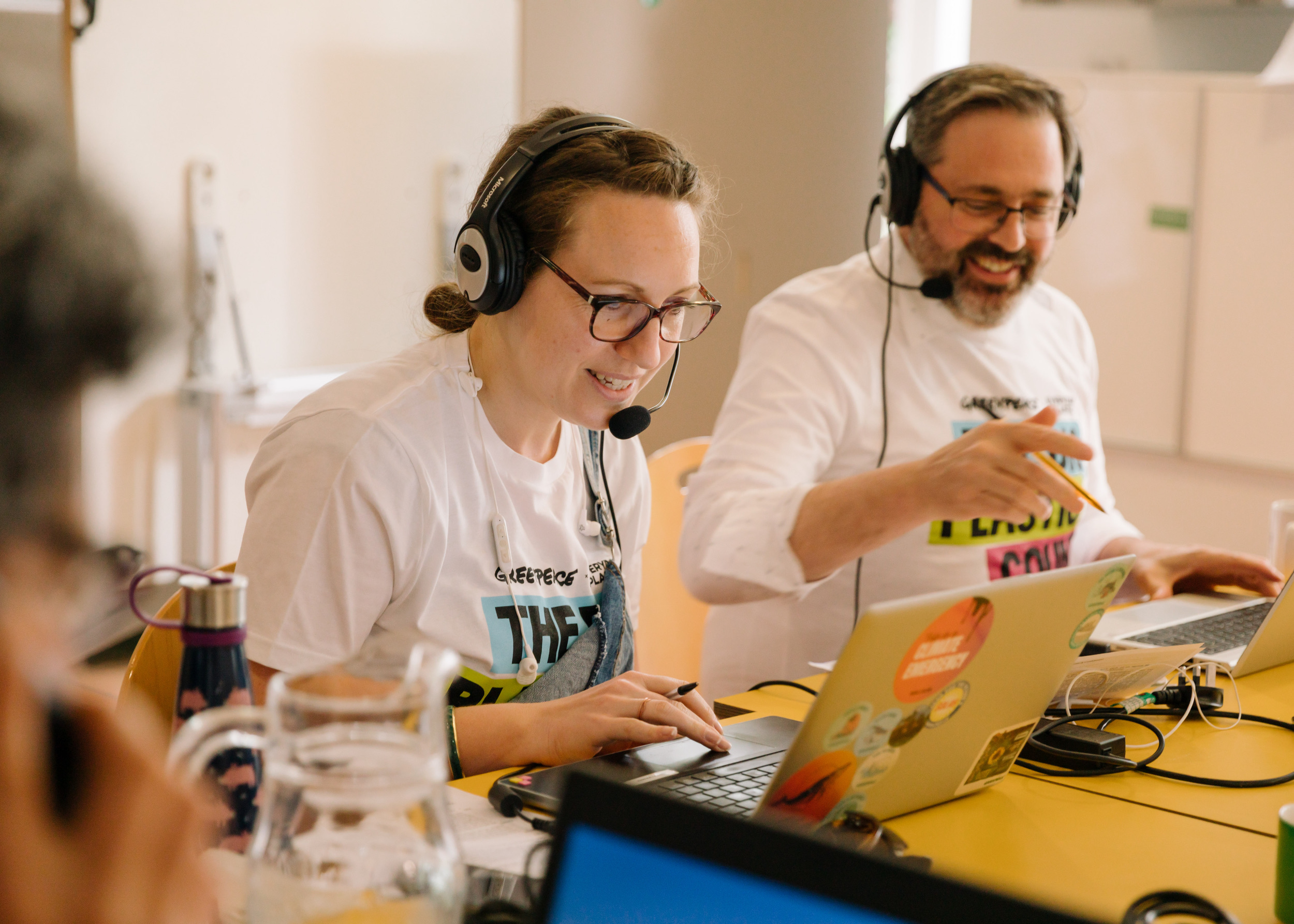 Two Greenpeace staff in matching t-shirts smile as they speak into headsets and look at laptops.