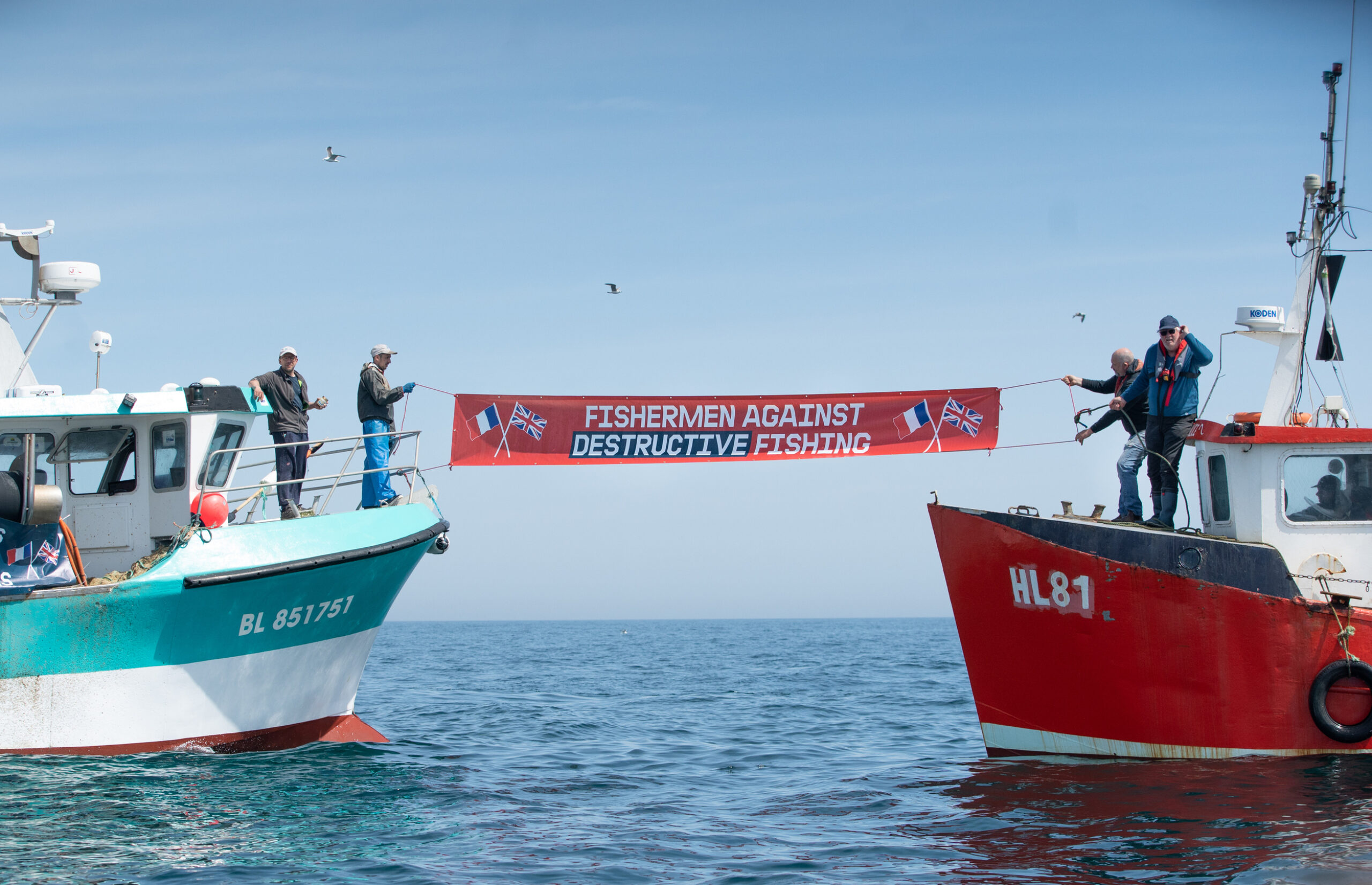 French and English fishermen in the middle of the water. They hold a sign between the two boats that reads "Fishermen against destructive fishing"