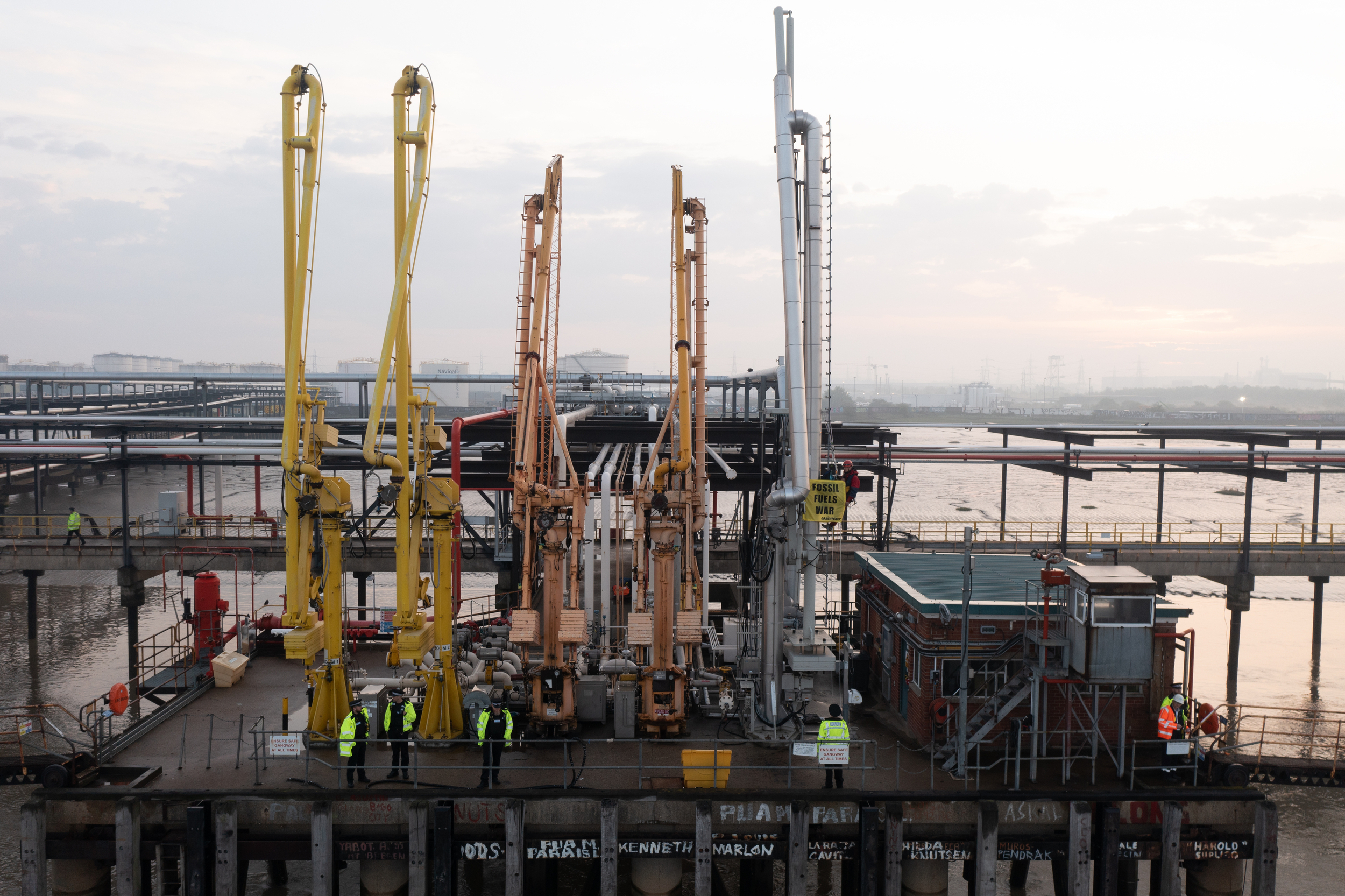 Tall pipe-like structures stretch towards the sky at an oil terminal by the sea where fuel tankers dock. An activist hangs from them flying a banner “Fossil fuels war”.