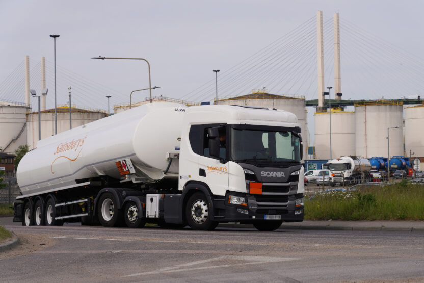 A tanker lorry with the Sainsburys logo on the side pulls away from a terminal complex. White industrial silos are visible in the background.