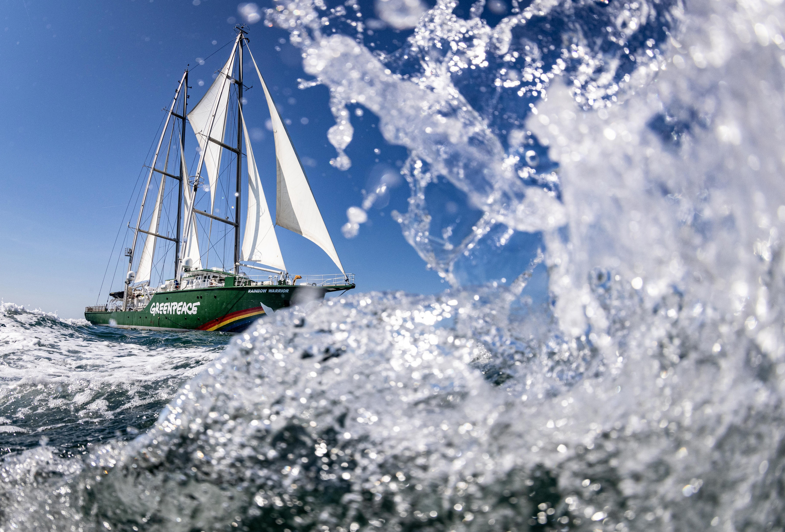 Water splashes in the foreground as a large ship sails along the sea in the background